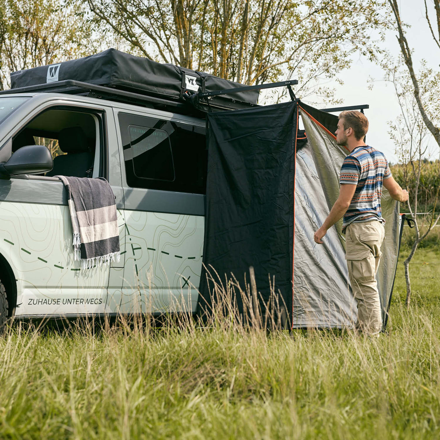 Person stellt schwarzes Vickywood Duschzelt mit Regenhaube neben weißem Van auf grüner Wiese auf.