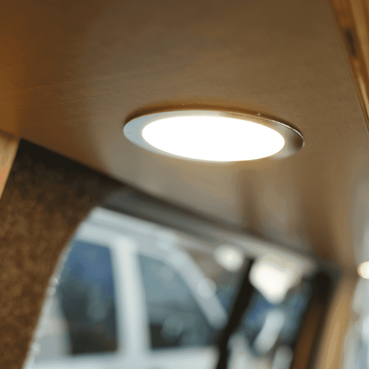 Runde LED-Einbauleuchte im Deckenspot mit warmweißem Licht, eingelassen in Holzdecke, darunter unscharf Fenster und Fahrzeug