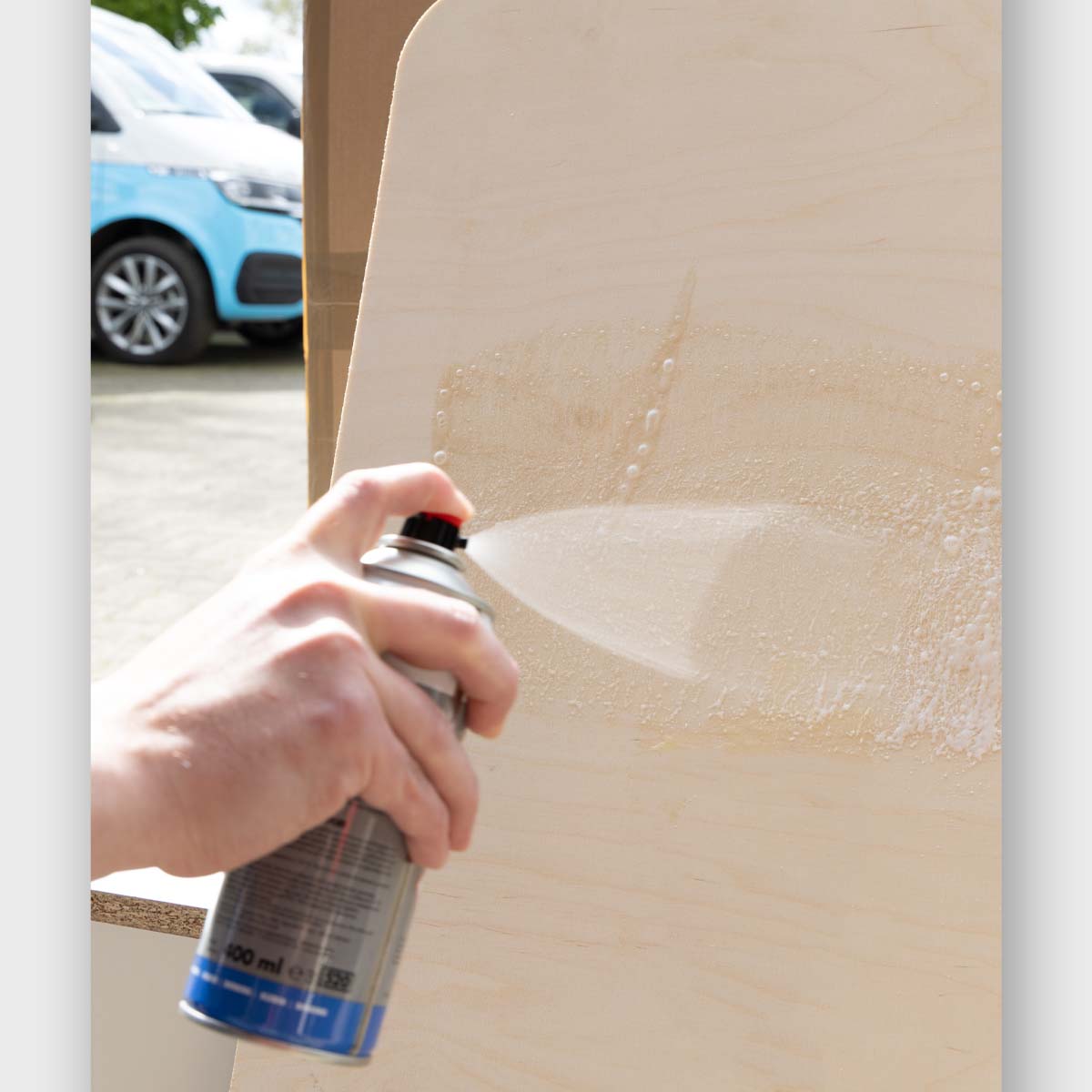 Hand spraying Kraft Sprühkleber Plus adhesive spray on wooden surface with visible mist from 400ml aerosol can
