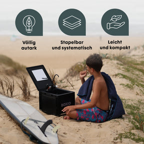 BOXIO WASH PLUS auf Sanddüne mit Surfbrett und sitzendem Jungen; Icons für autark, stapelbar, leicht und kompakt sichtbar