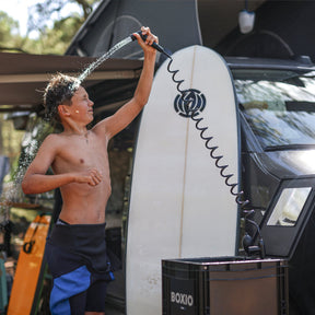 Junge duscht mit BOXIO WASH PLUS neben Surfbrett am Van; mobiler Wasserhahn mit Spiralschlauch und Wasserstrahl sichtbar