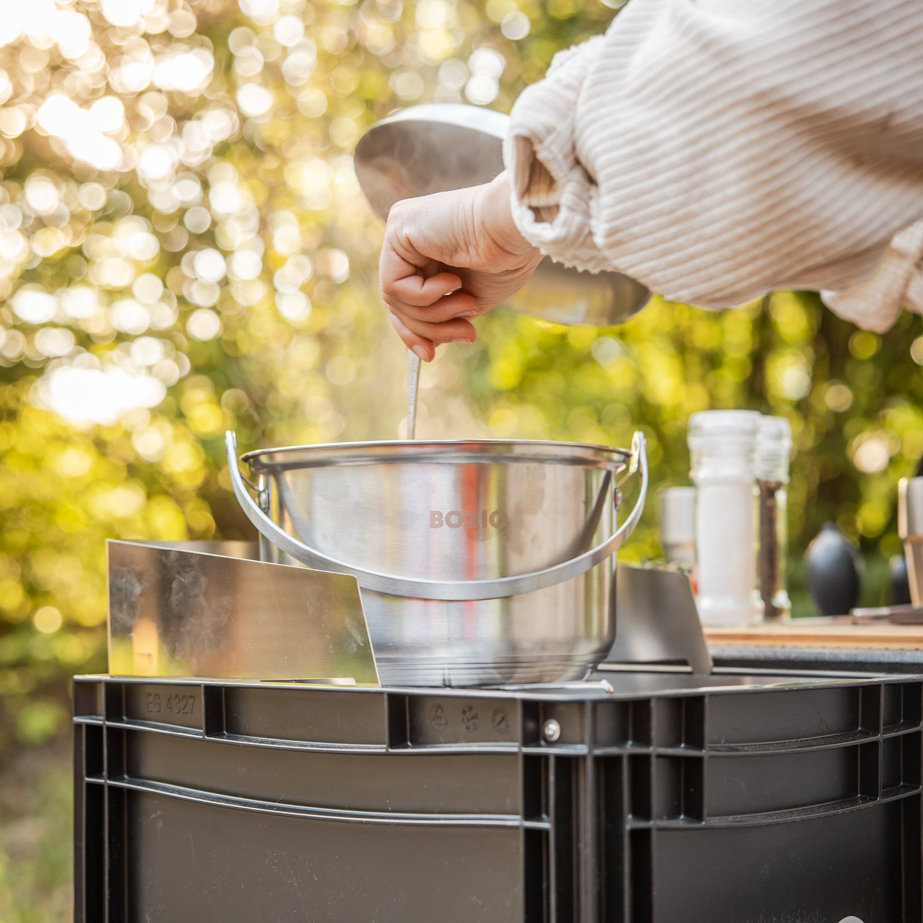 Camping-Kochset BOXIO COOK MAX+ mit Edelstahltopf auf schwarzer Box; Hand rührt am Outdoor-Kocher, Dampf steigt auf