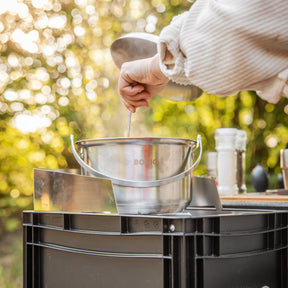 Camping-Kochset BOXIO COOK MAX+ mit Edelstahltopf auf schwarzer Box; Hand rührt am Outdoor-Kocher, Dampf steigt auf