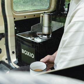 BOXIO COOK Gaskocher im Van mit Espressokanne und Emailletasse, Person sitzt daneben und hält Kaffee in der Hand