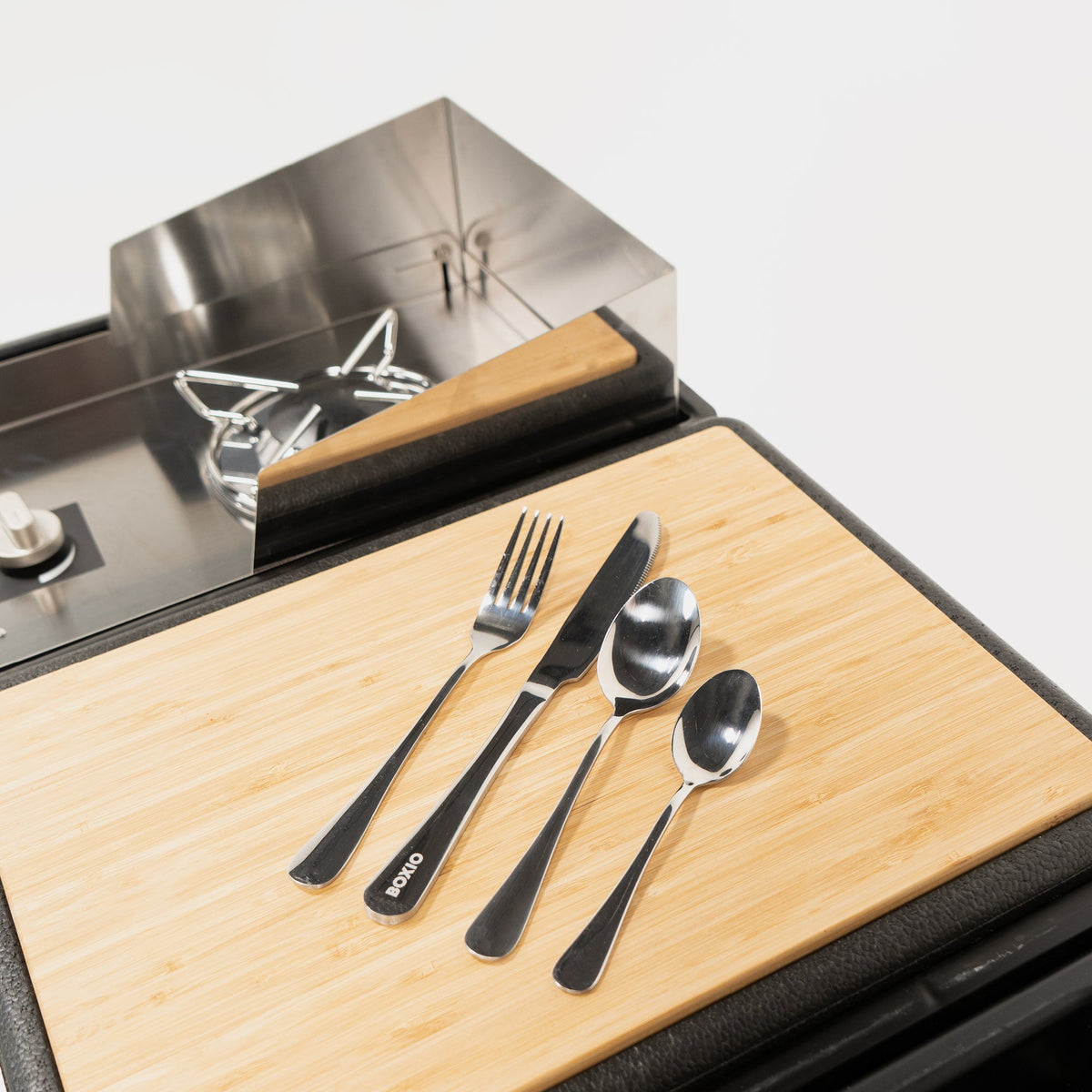 Camping-Besteck BOXIO EAT mit Gabel, Messer und zwei Löffeln auf hellem Bambusbrett neben Metallbox und Deckelglocke