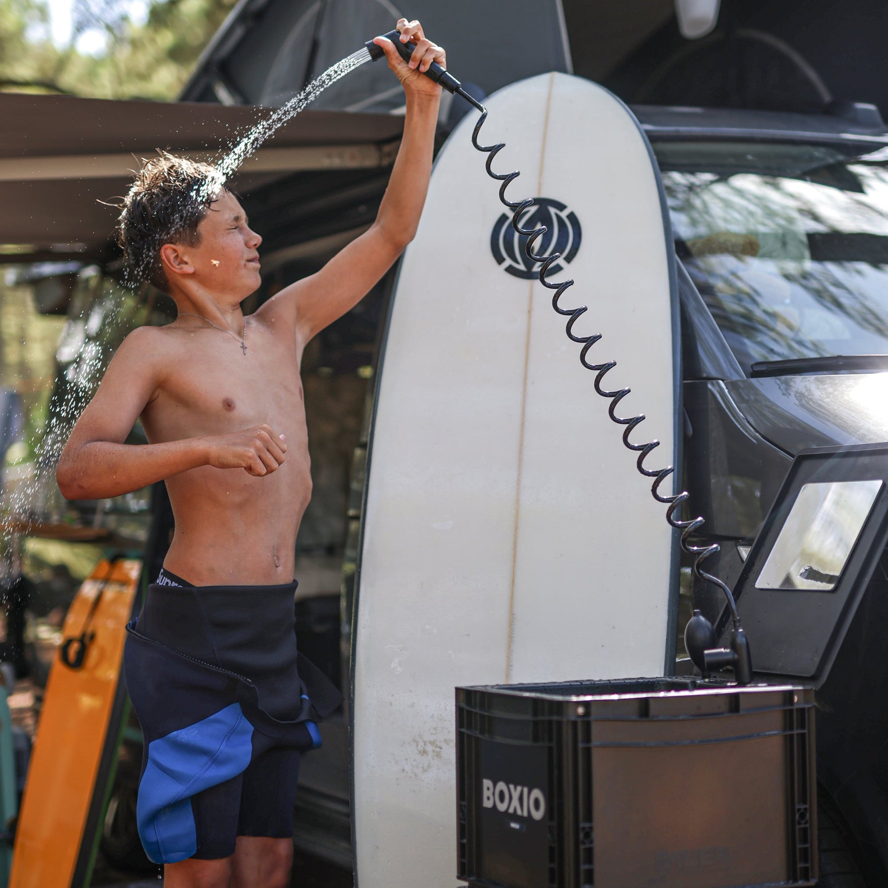 Junge duscht draußen am Wohnmobil mit BOXIO SANITARY PLUS und Surfbrett daneben, Wasserstrahl trifft Kopf und Oberkörper