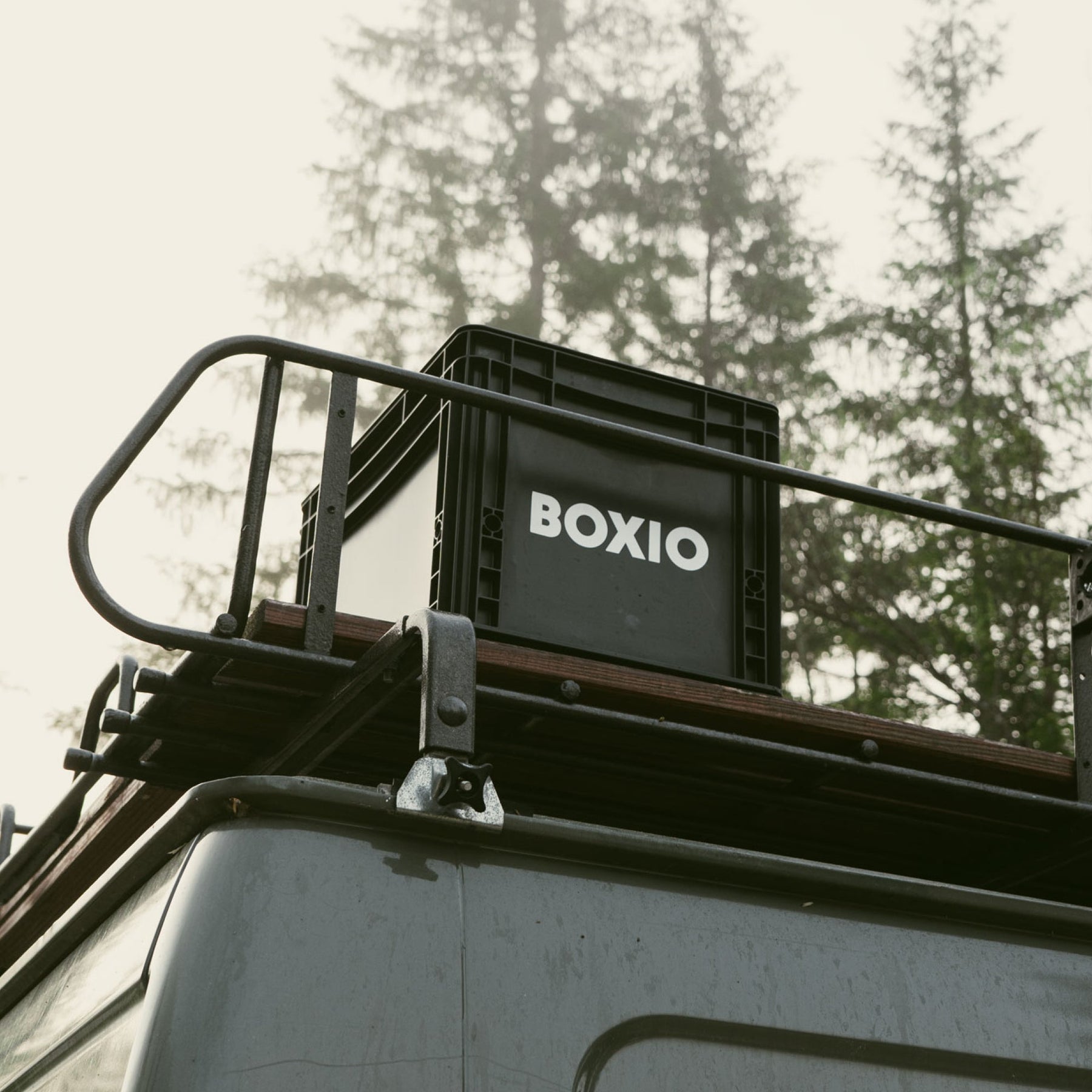 BOXIO SOLO Eurobox mit Deckel auf Dachgepäckträger eines Fahrzeugs, schwarze Box mit weißem Logo vor Waldhintergrund