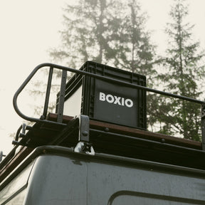BOXIO SOLO Eurobox mit Deckel auf Dachgepäckträger eines Fahrzeugs, schwarze Box mit weißem Logo vor Waldhintergrund