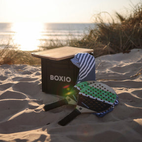 BOXIO SOLO Eurobox mit Deckel auf Sandstrand neben Handtuch und Sandever Strandschlägern bei Sonnenuntergang am Meer