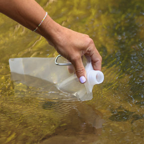Hand mit Armband hält BOXIO WASH UP Eurobox-Wasserfilter im klaren Wasser; transparenter Behälter mit Schrauböffnung sichtbar