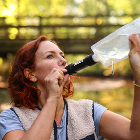 Frau trinkt am BOXIO WATER FILTER aus transparentem Beutel im Outdoor-Einsatz, rotes Haar, Fleeceweste, Waldhintergrund