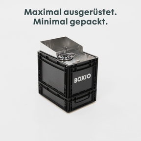 BOXIO COOK MAX+ Camping-Kochset aus schwarzer Kiste mit Edelstahlkocher und Windschutz; darüber Schriftzug „Maximal ausgerüstet. Minimal gepackt.“
